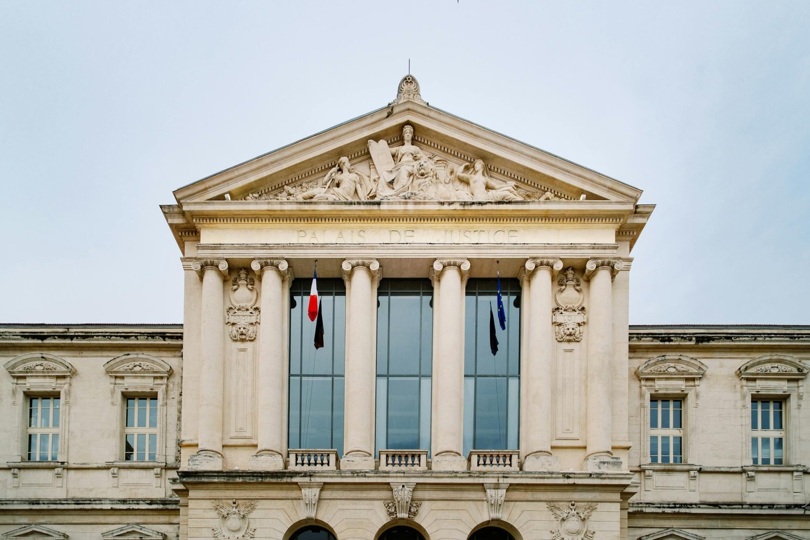 Façade d’un palais de justice (abritant le tribunal correctionnel) de style néoclassique, avec colonnes imposantes, fronton sculpté et inscription « Palais de Justice », surmonté de statues allégoriques, avec drapeaux français et européen devant de grandes fenêtres.